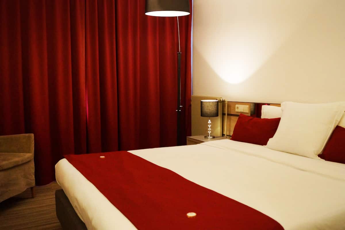 A hotel room with a large bed, red curtains, a nightstand with a lamp, and a chair.