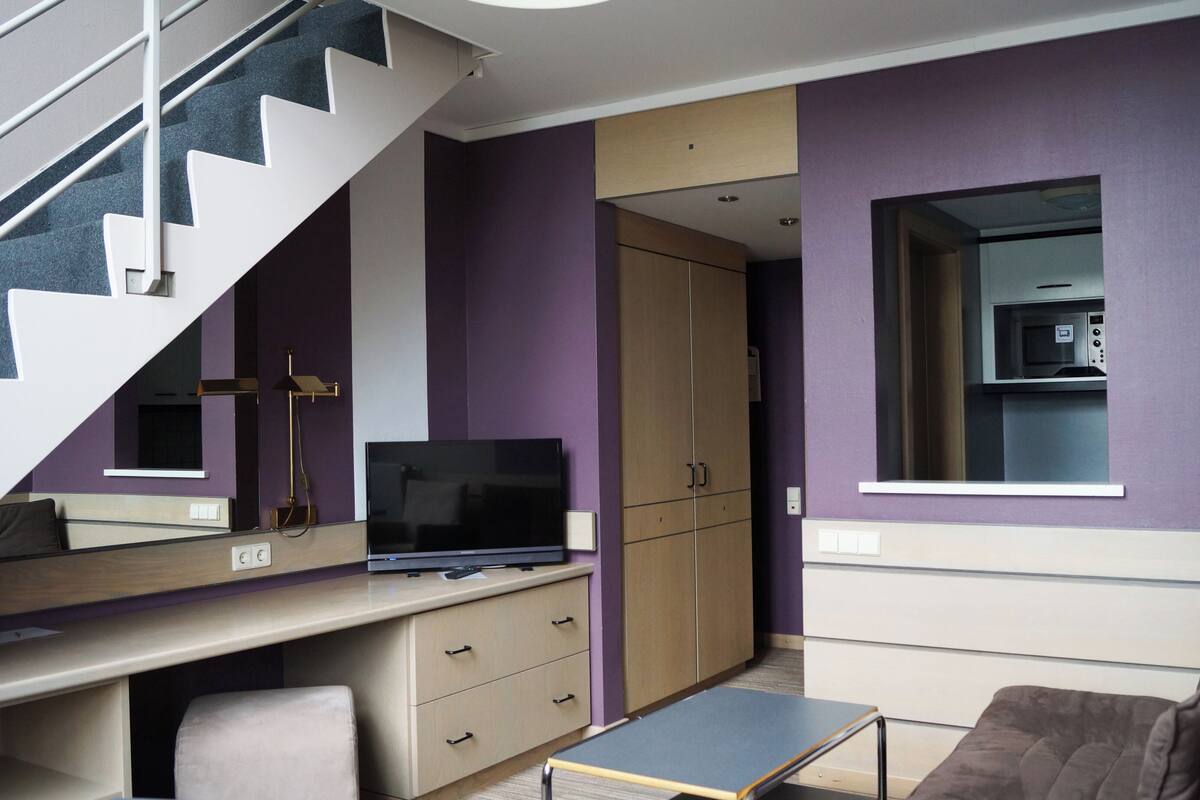 A modern room with a desk, a television, a sofa, a staircase, and a wardrobe.