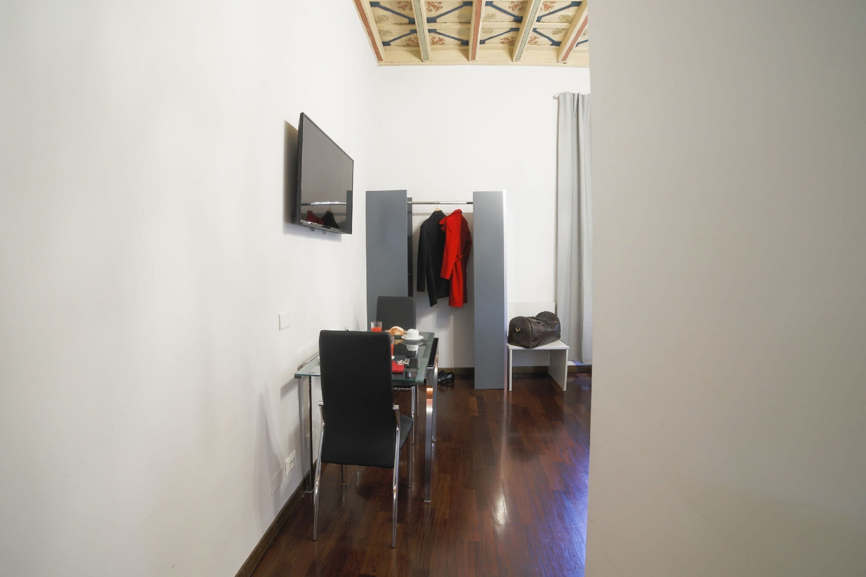 A compact room with a wooden floor, a dining table with chairs, a coat rack with hanging clothes, a mirror, and a television mounted on the wall.