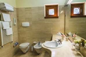 Un baño con inodoro, bidé, lavabo, toallero y dos ventanas pequeñas con marcos de madera.