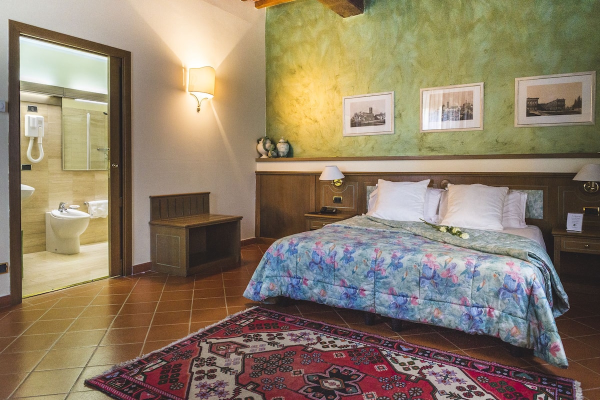 Hotelkamer met een bed, nachtkastje, vloerkleed en een badkamer zichtbaar door een geopende deur.