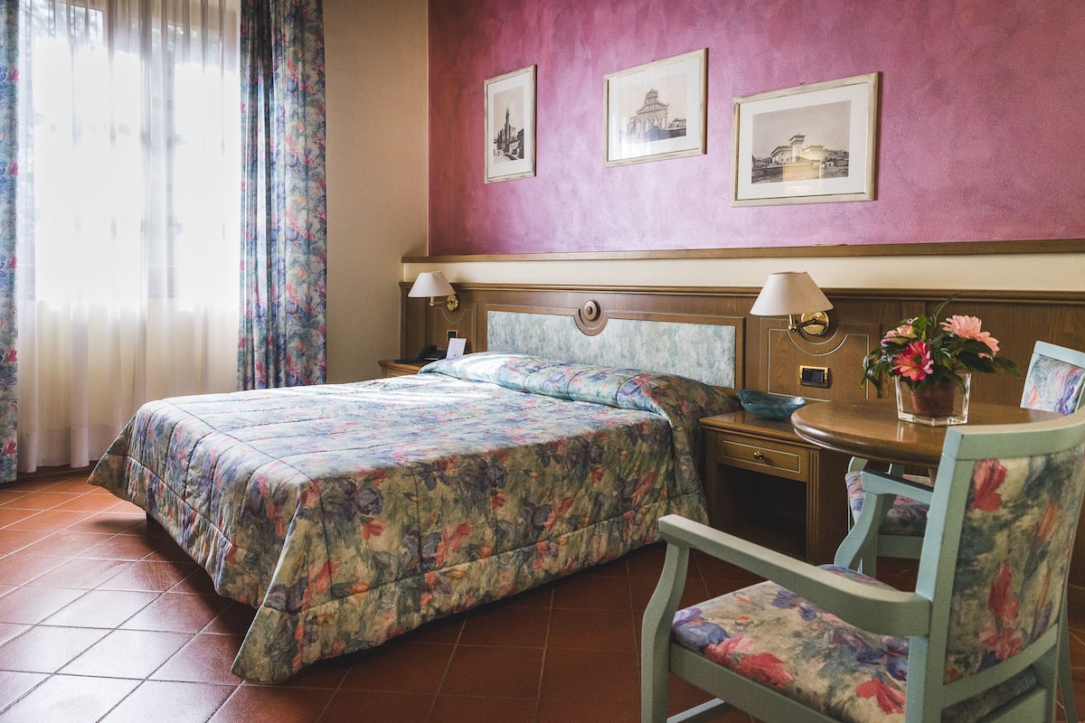 Een hotelkamer met een bed, een stoel, een tafel met een vaas en twee ingelijste foto's aan de muur.