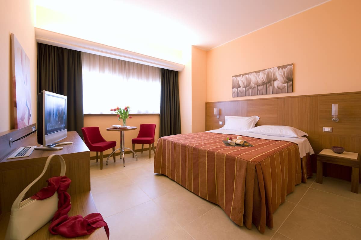 A hotel room with a bed, a desk with a computer, a chair, a small table, and a window with curtains.