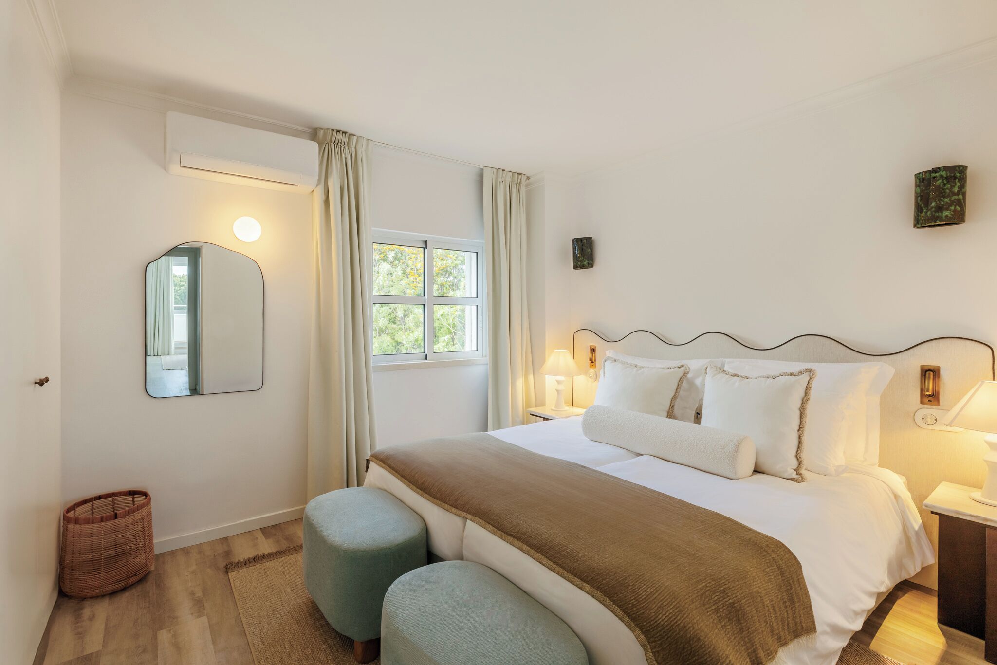 A hotel room with a bed, a mirror, a window, a lamp, and a wall-mounted air conditioner.