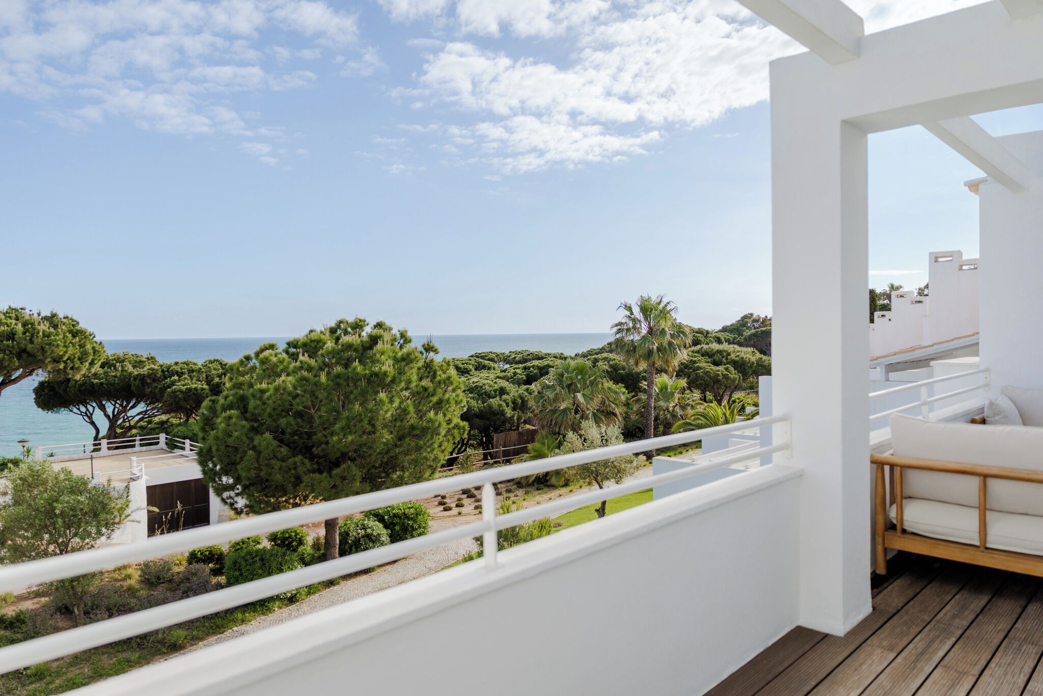 A balcony with a white railing, a wooden deck, and a view of the sea and greenery.
