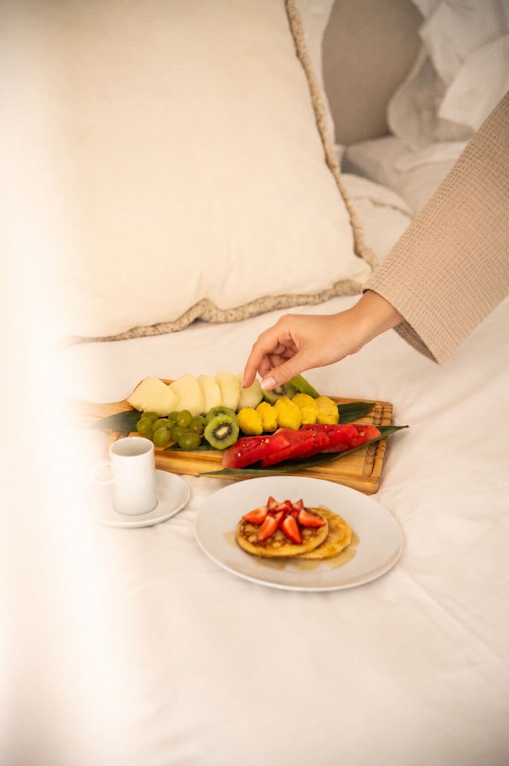 A breakfast setting with a plate of pancakes topped with strawberries, a bowl of fruit including kiwi and watermelon, and a cup of coffee on a bed.