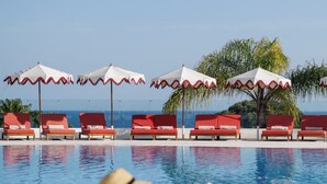 A pool area with lounge chairs and umbrellas, a person sitting by the pool, and a clear view of the ocean. - Masana Algarve, a Destination by Hyatt (Albufeira)