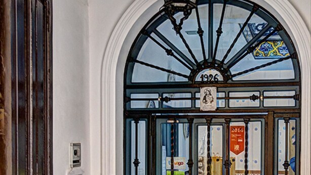 A door with a blue and white patterned tile design, a black metal gate, and a decorative light fixture above it. - Pensión Montoreña (Seville)