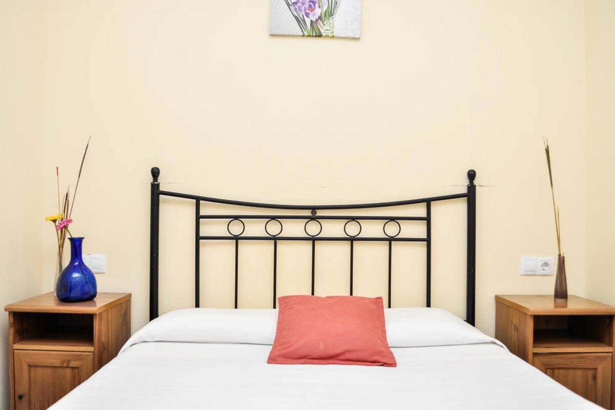 A neatly made bed with a red pillow, a black metal headboard, and two wooden nightstands with vases.