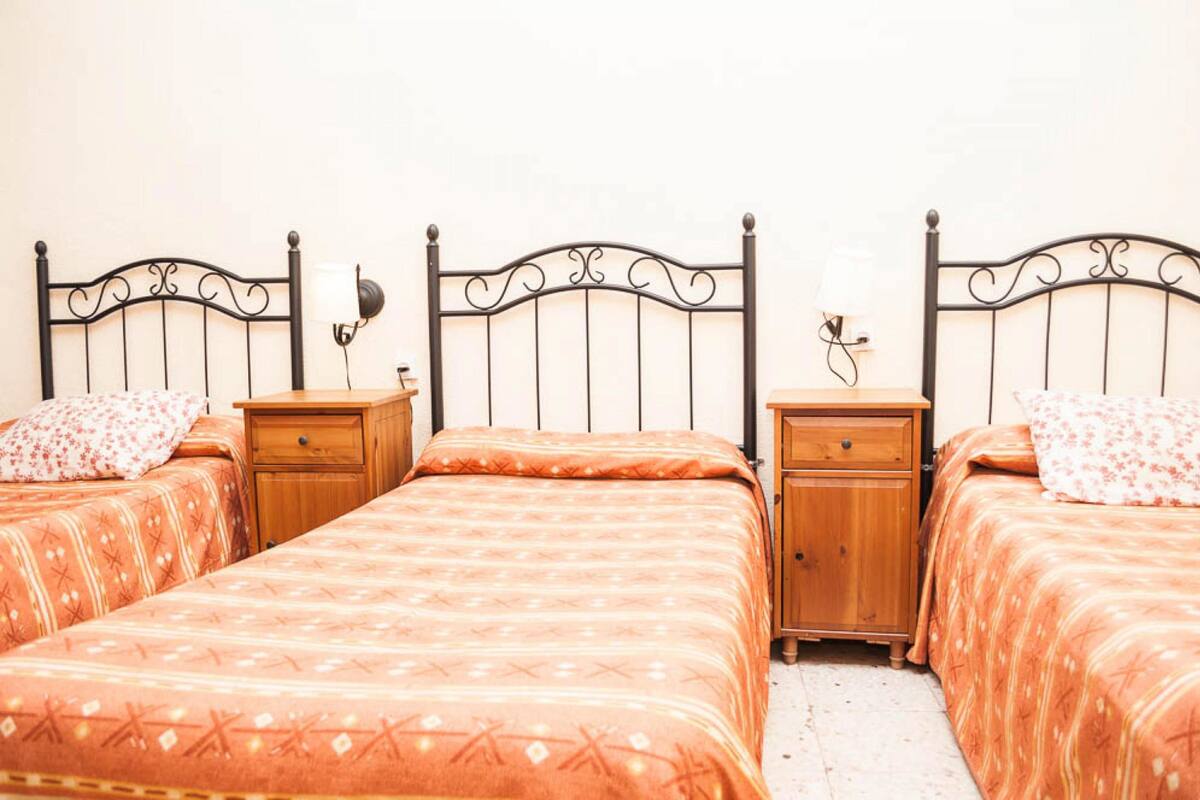 A room with three beds aligned against a wall, each with a wooden nightstand and a lamp.