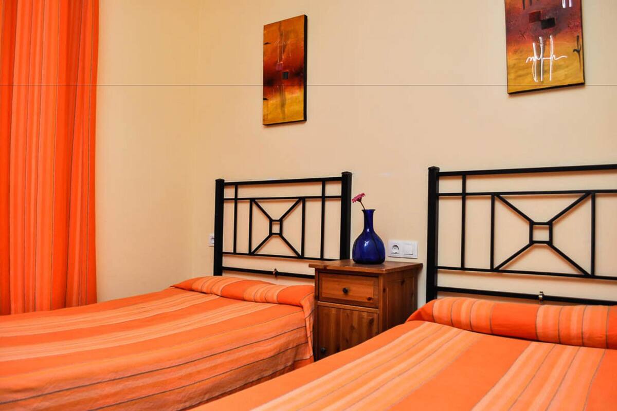 A hotel room with two beds, orange bedding, a wooden nightstand, and two wall-mounted paintings.