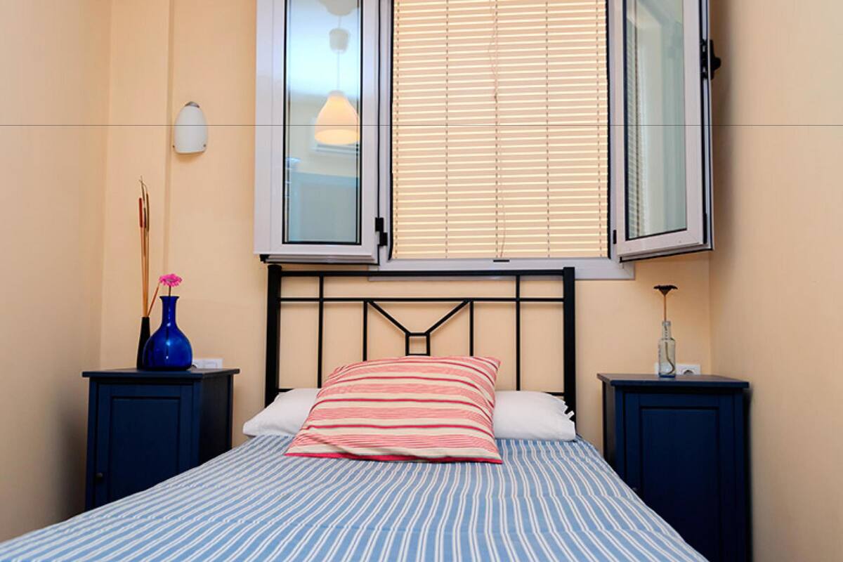 A bedroom with a bed, two nightstands, a window with blinds, a vase, and a lamp.