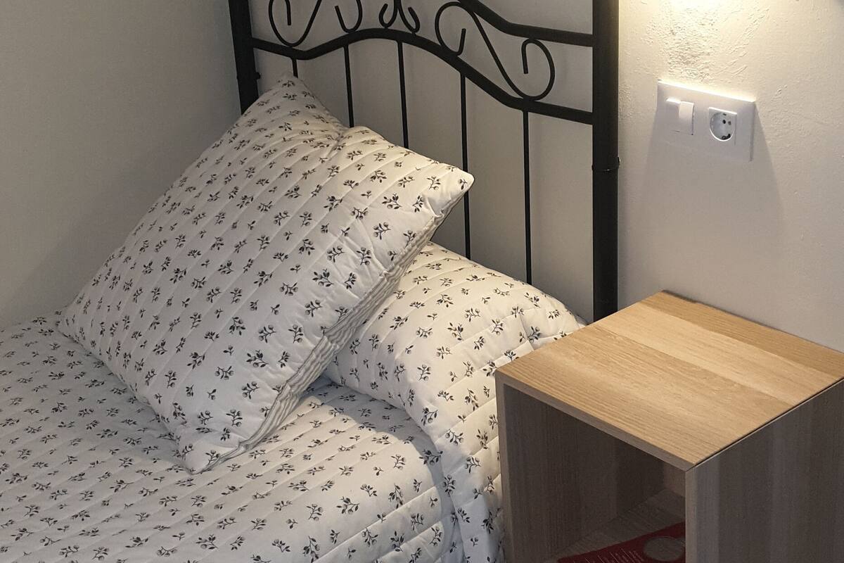 A bed with a floral-patterned bedspread and pillows, a wooden bedside table, and a wall-mounted light fixture.