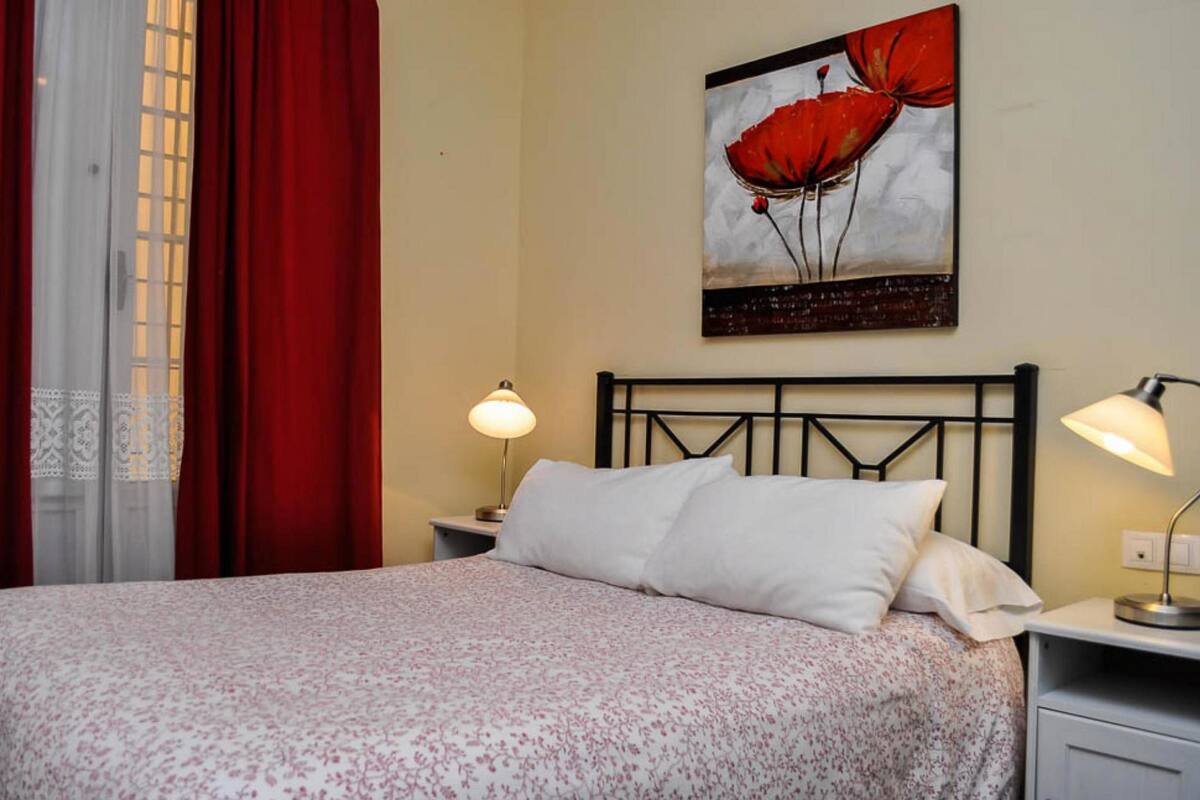 A neatly made bed with a floral bedspread, two bedside lamps, and a painting of red flowers on the wall.