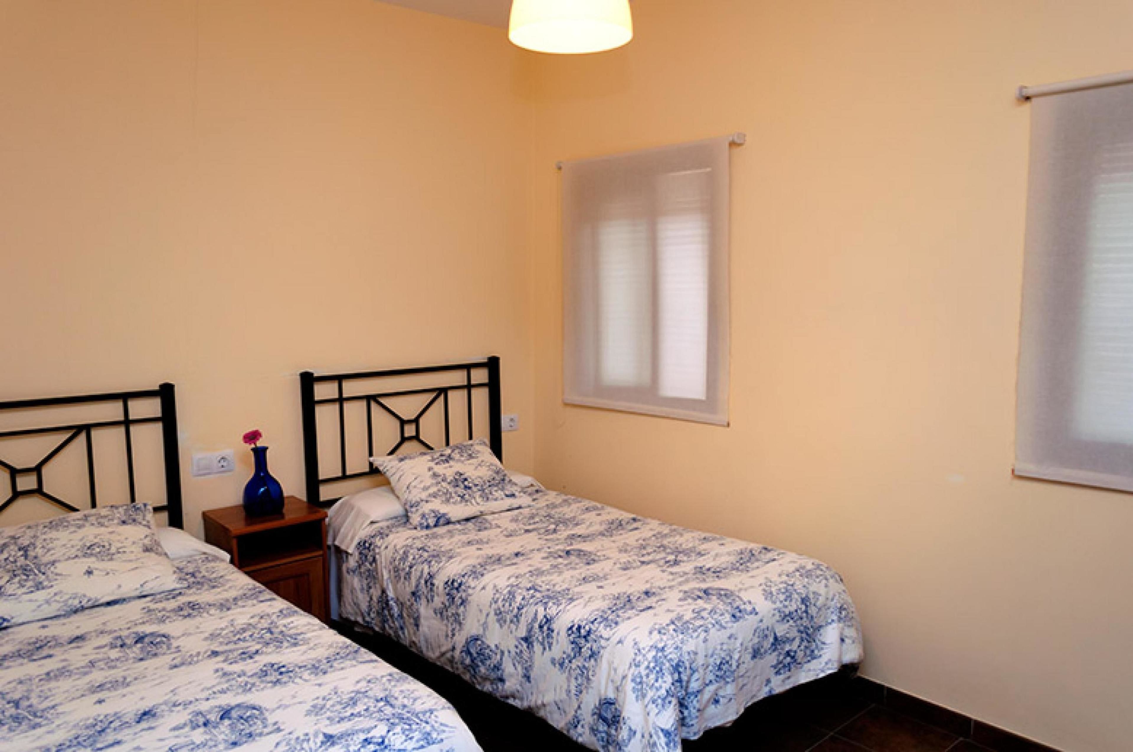 Een slaapkamer met twee eenpersoonsbedden, een nachtkastje, een vaas en een raam met jaloezieën.