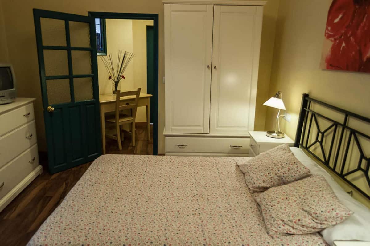 A bedroom with a bed, a desk, a chair, a wardrobe, and a nightstand with a lamp.