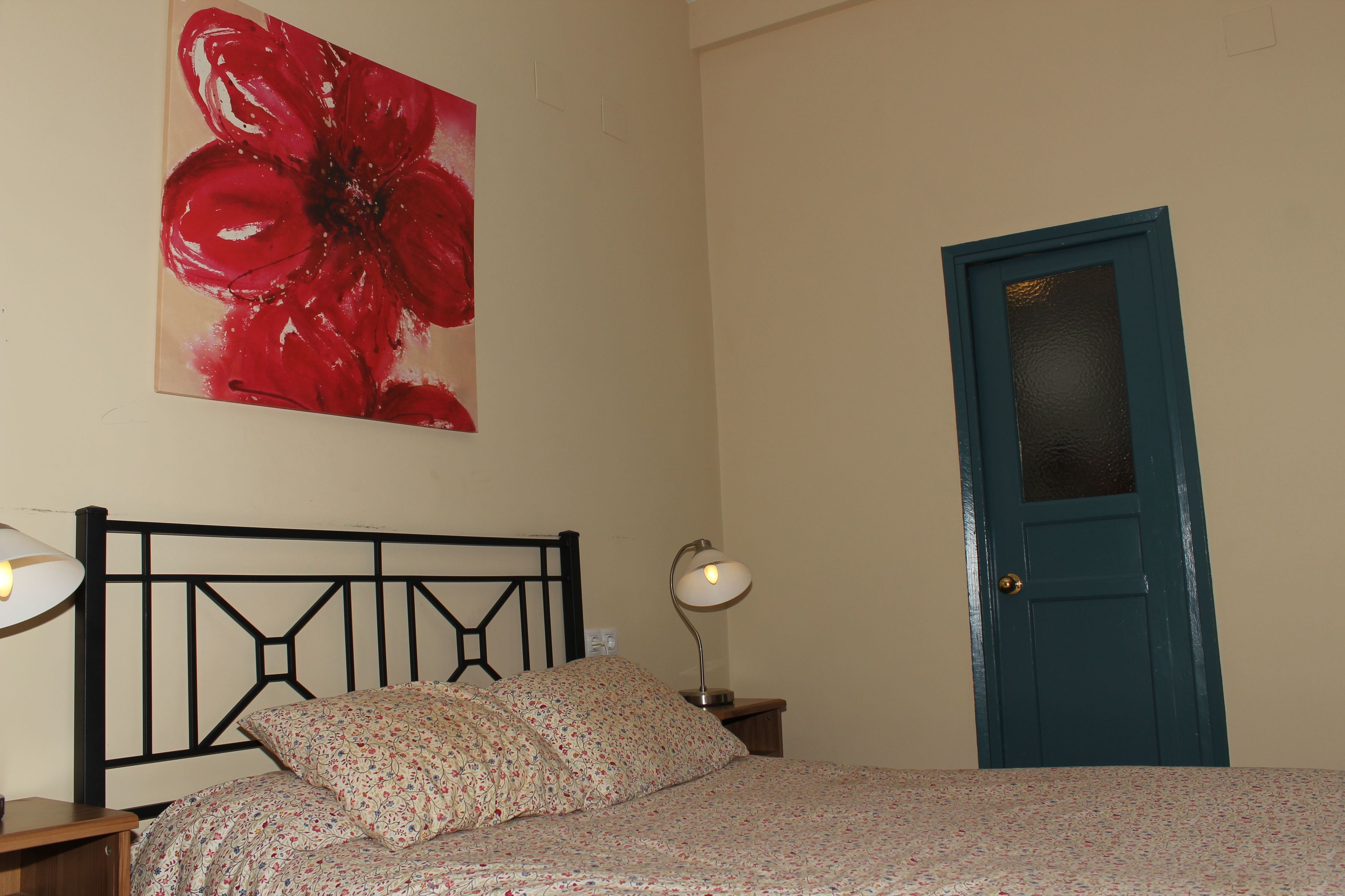 A bedroom with a bed, a nightstand, a lamp, a door, and a floral painting on the wall.
