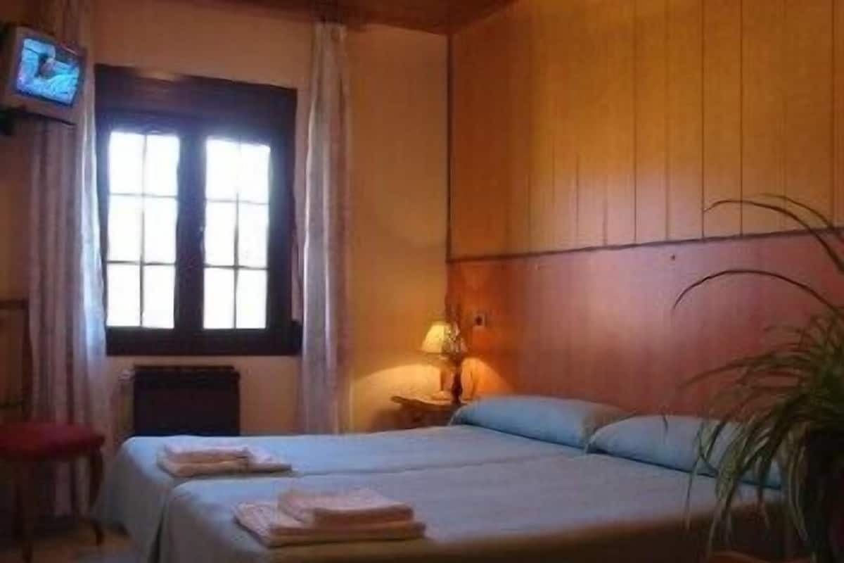 A room with two beds, a television, a window with curtains, and a bedside table with a lamp.