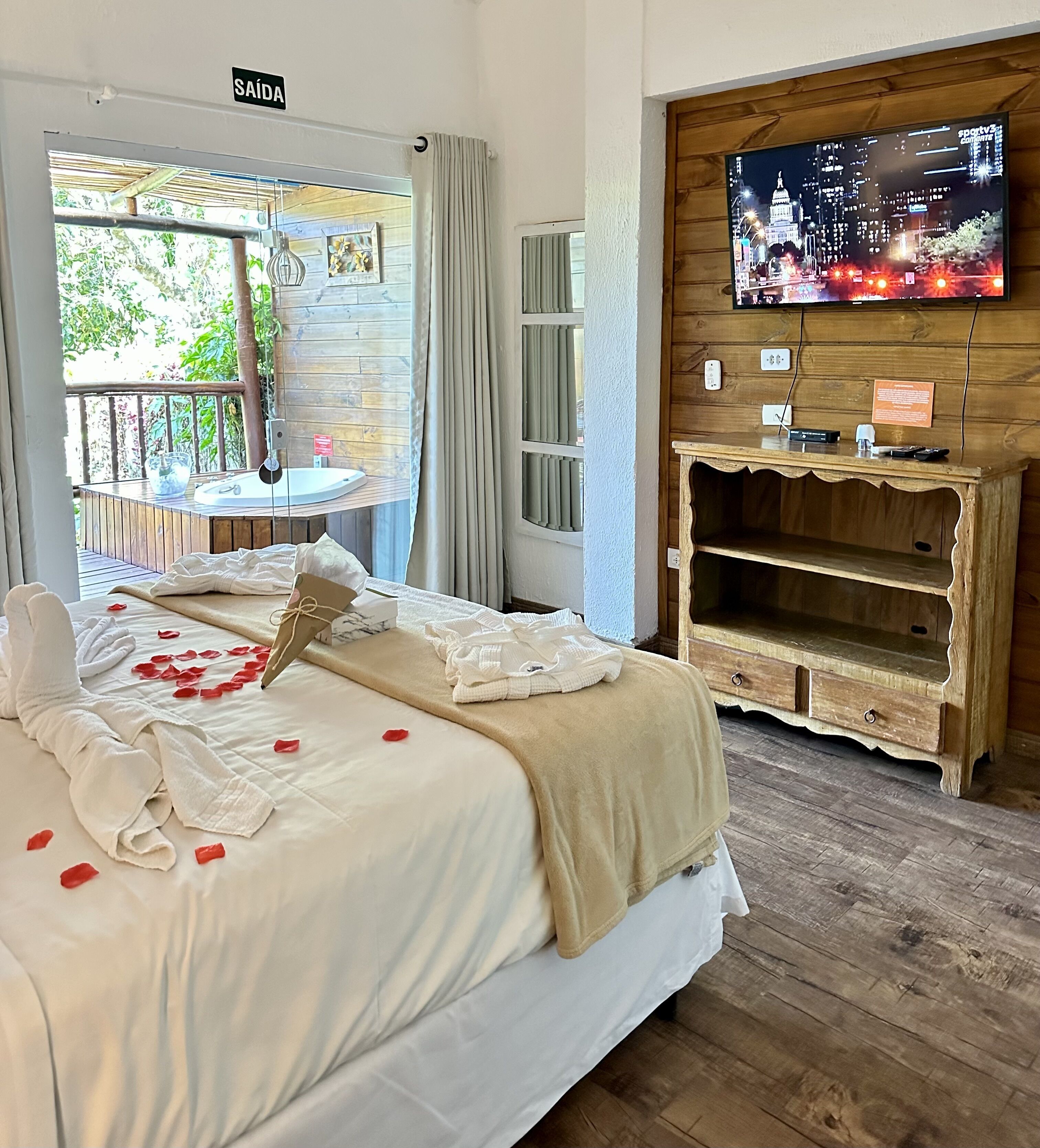 Quarto de hotel com cama, TV numa parede de madeira e varanda com vista para o verde.