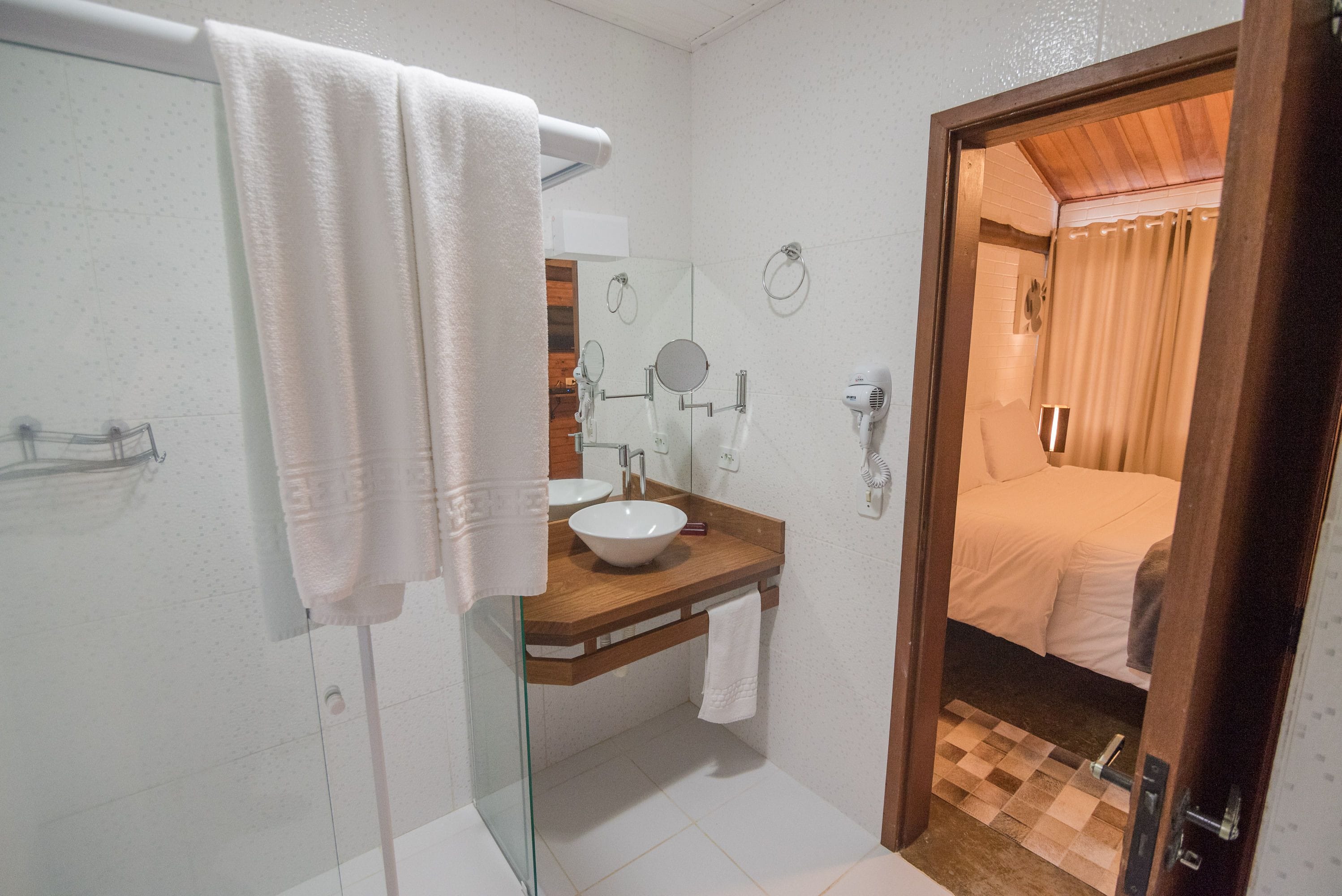 A bathroom with a glass shower enclosure, a wooden vanity with a white sink, and a visible bed in the adjacent room.