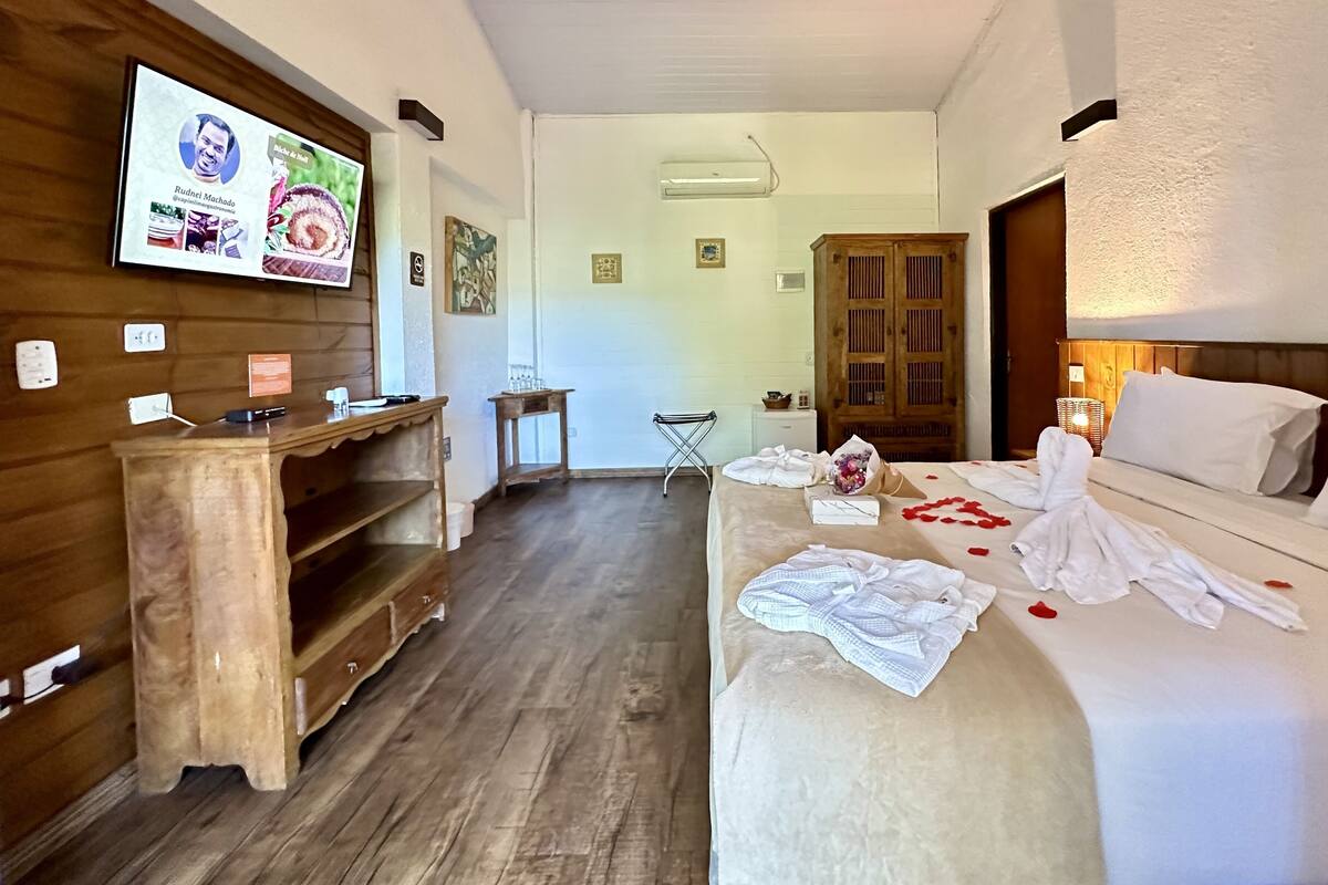 A hotel room with a wooden desk, a television displaying a menu, a bed with rose petals, and a wooden wardrobe.