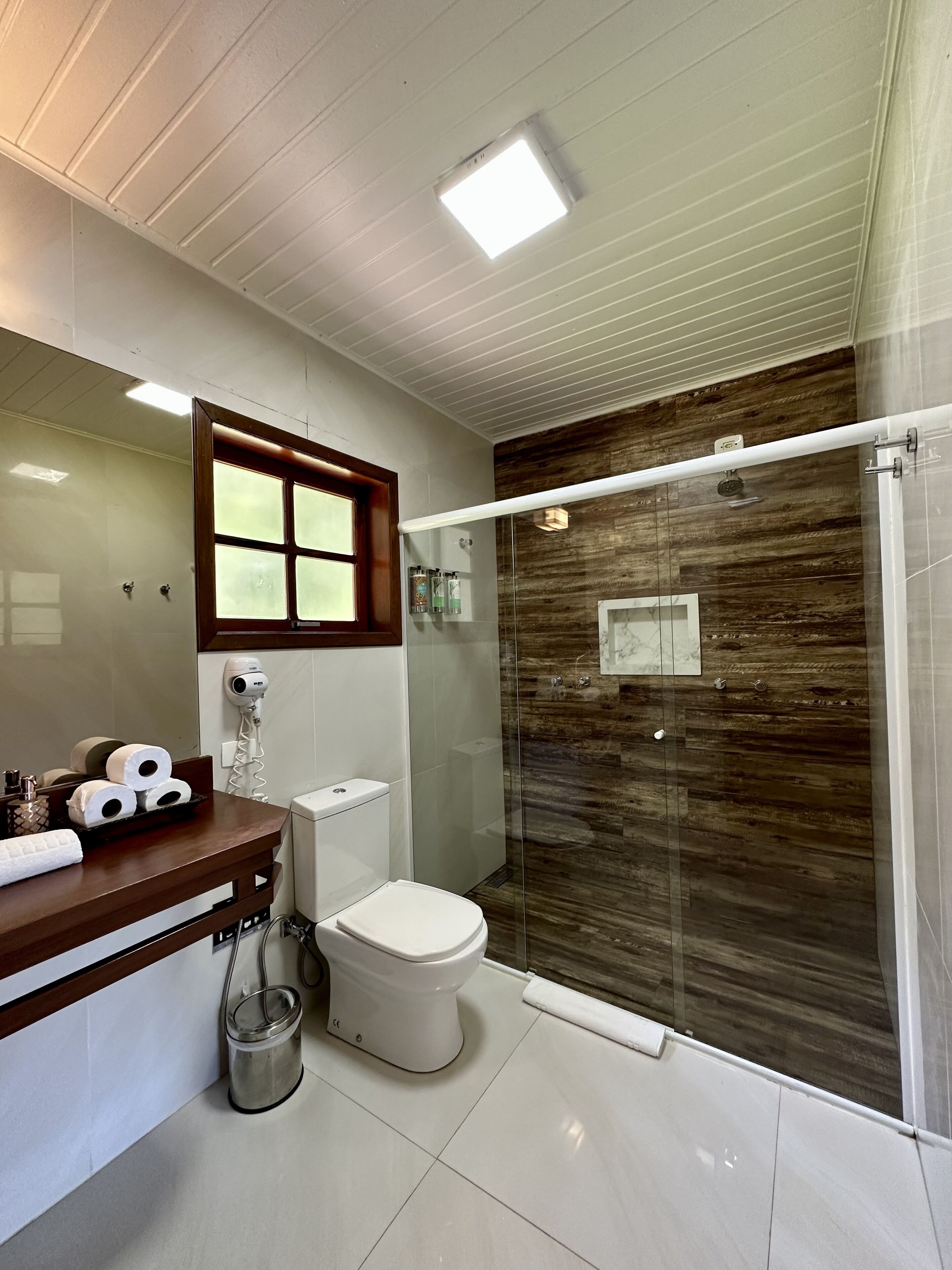 Un baño moderno con ducha de vidrio, inodoro, lavabo con encimera de madera y espejo con ventana arriba.
