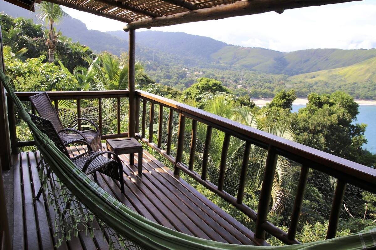 A wooden deck with a hammock, chairs, and a small table overlooking a lush landscape with mountains and a body of water.