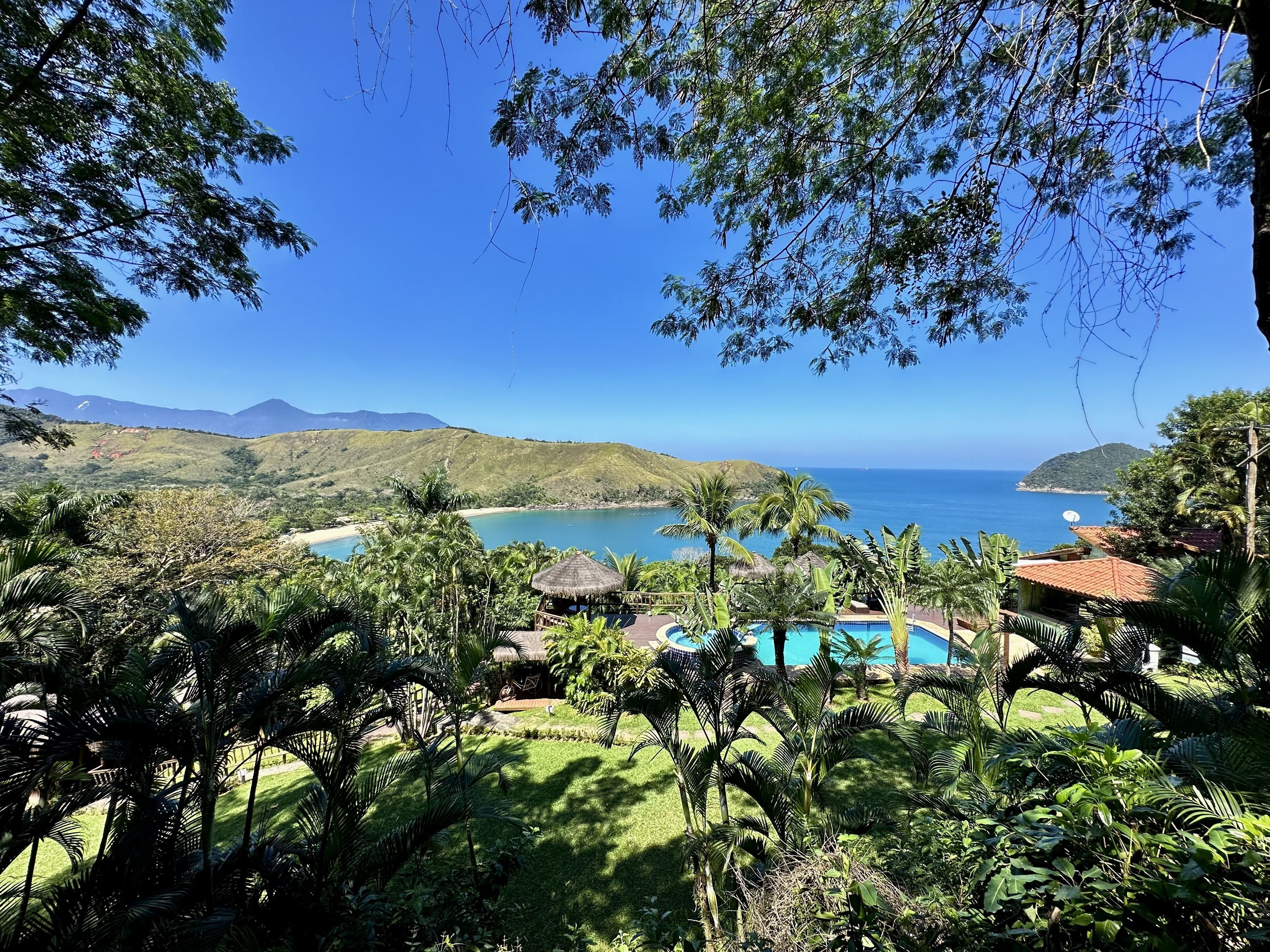 A tropical resort with a swimming pool, lush greenery, and a view of the ocean and distant mountains.