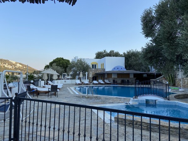 A pool area with lounge chairs and umbrellas, a stone building with a yellow door, and a view of a hillside.