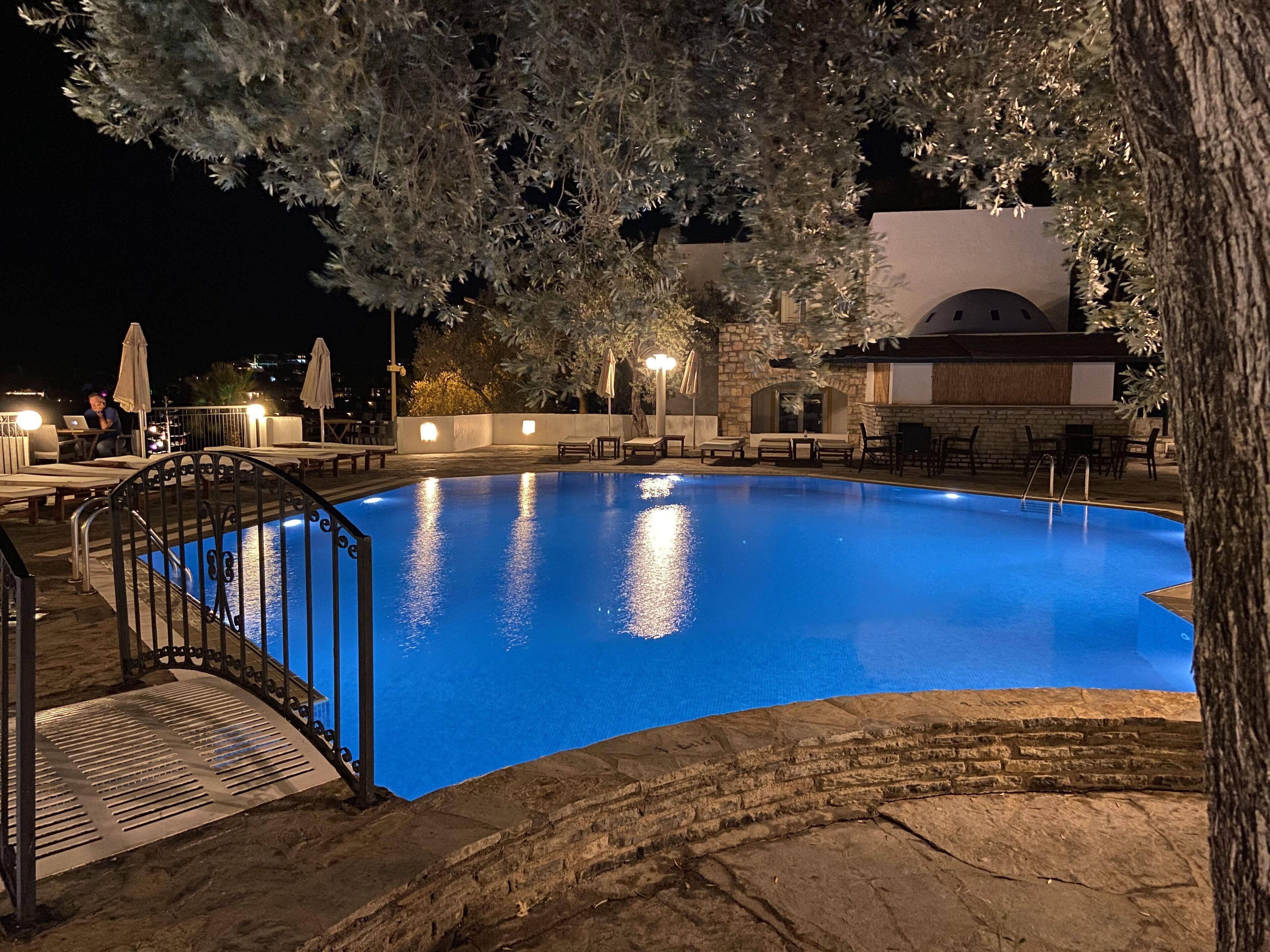 Una piscina nocturna con terraza de piedra, sillas de descanso y sombrillas.