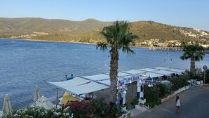 Eine Küstenlandschaft mit Restaurant direkt am Strand, Palmen und Blick auf das Meer und die dahinterliegenden Hügel.