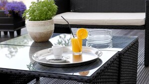 Una mesa de vidrio con una bandeja, un vaso de jugo de naranja y una taza blanca sobre una silla de mimbre.