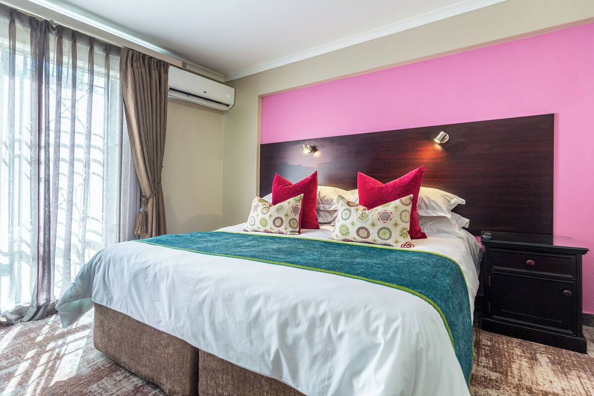 A hotel room with a large bed, a headboard with a light fixture, a nightstand, and a window with curtains.