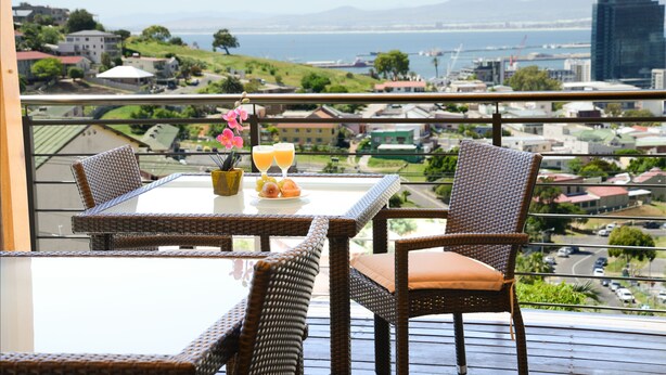 A balcony with wicker furniture, a table with a glass top, and a view of a coastal cityscape. - Signal Hill Lodge (Cape Town)