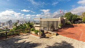 A paved pathway leading to a building with a metal fence and flags, surrounded by greenery and a cityscape in the background. - Signal Hill Lodge (Cape Town)