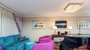 A living room with a teal sofa, a pink armchair, a blue ottoman, a black piano, and a flat-screen TV. - Signal Hill Lodge (Cape Town)