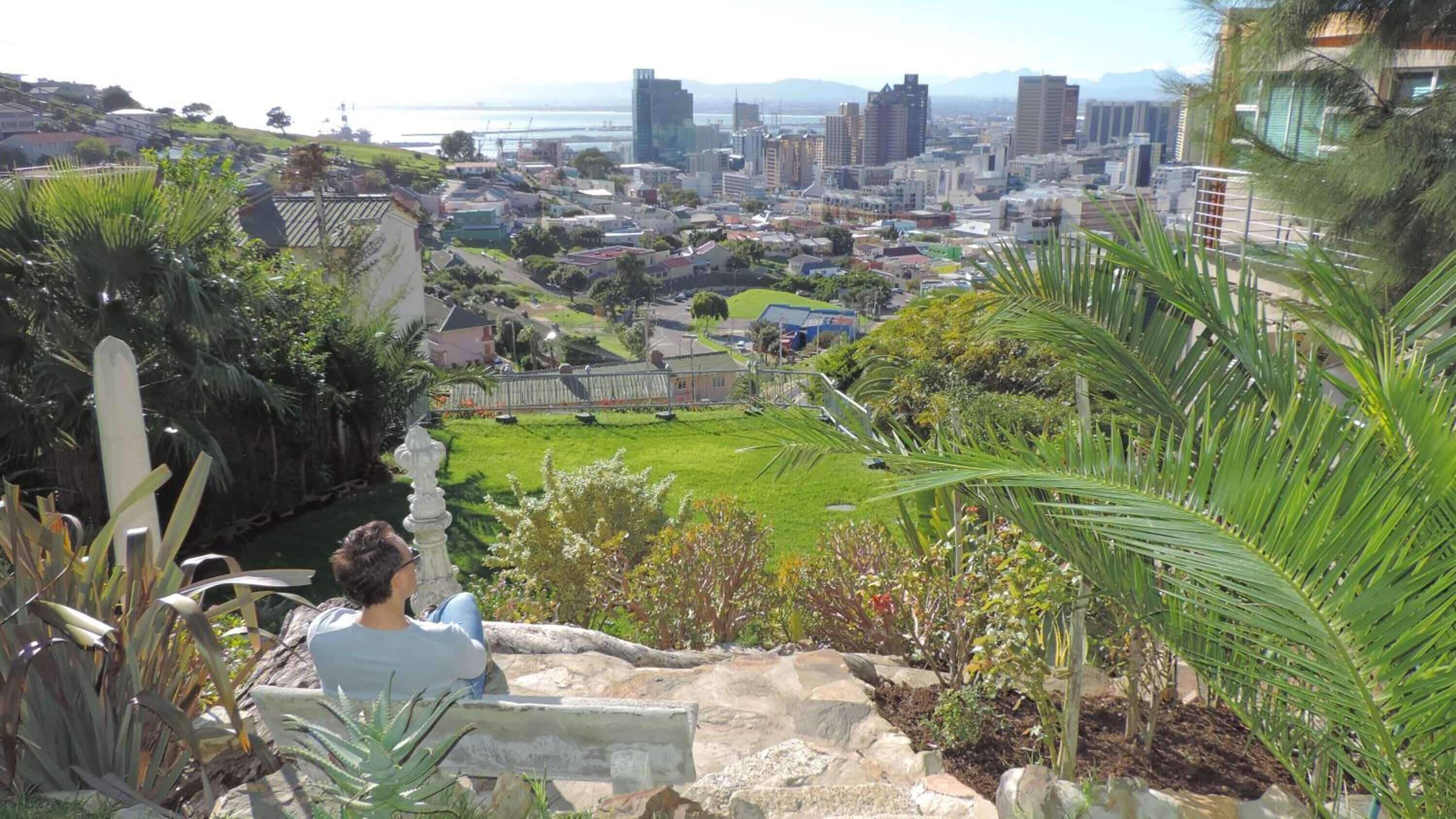 A person sitting on a stone bench overlooking a cityscape with a mix of residential and commercial buildings, a green park, and a body of water in the distance.