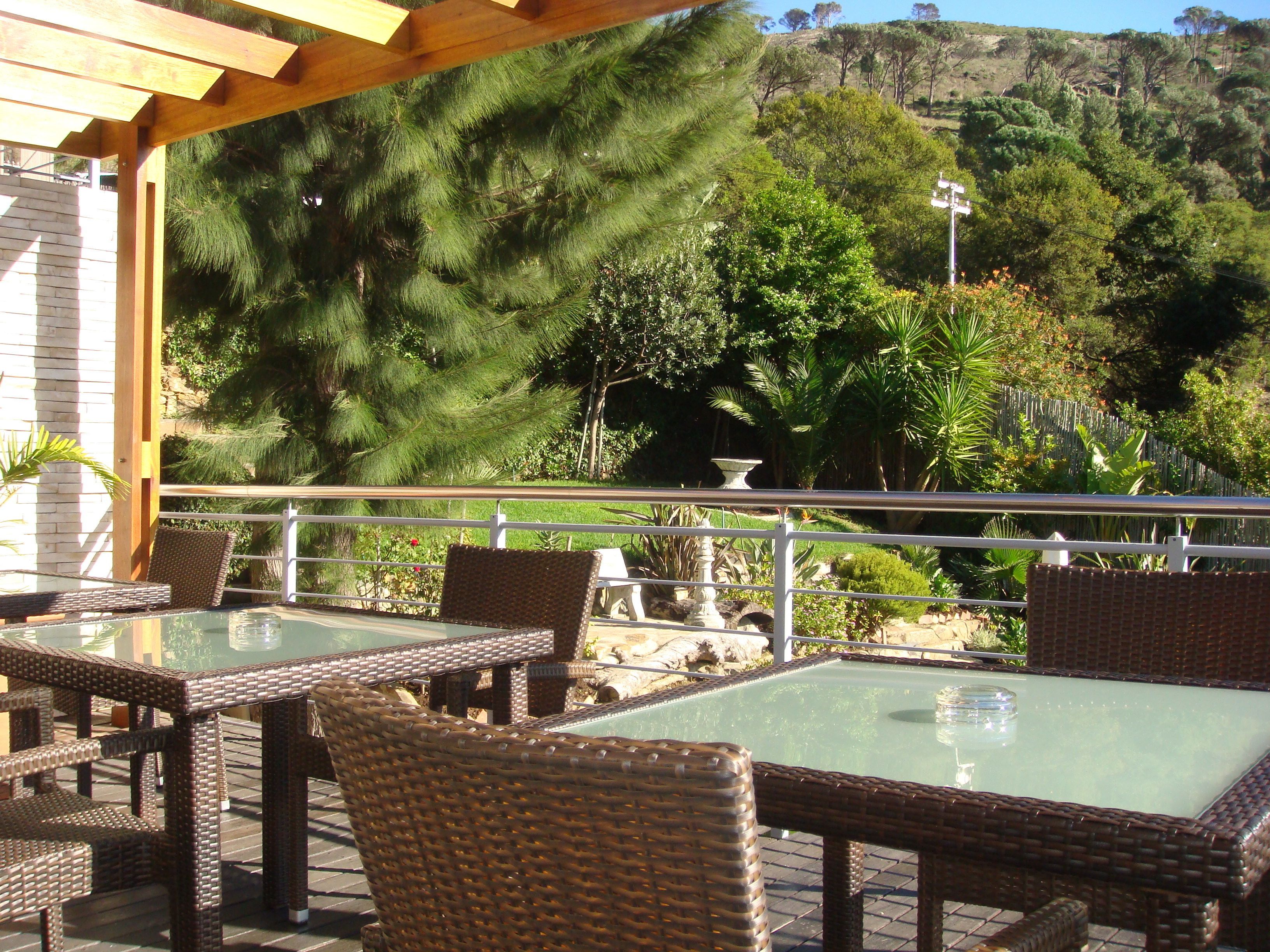 Un patio al aire libre con mobiliario de mimbre y una mesa con cubierta de vidrio, con vista a un jardín ajardinado con diversas plantas y una vista de una colina.
