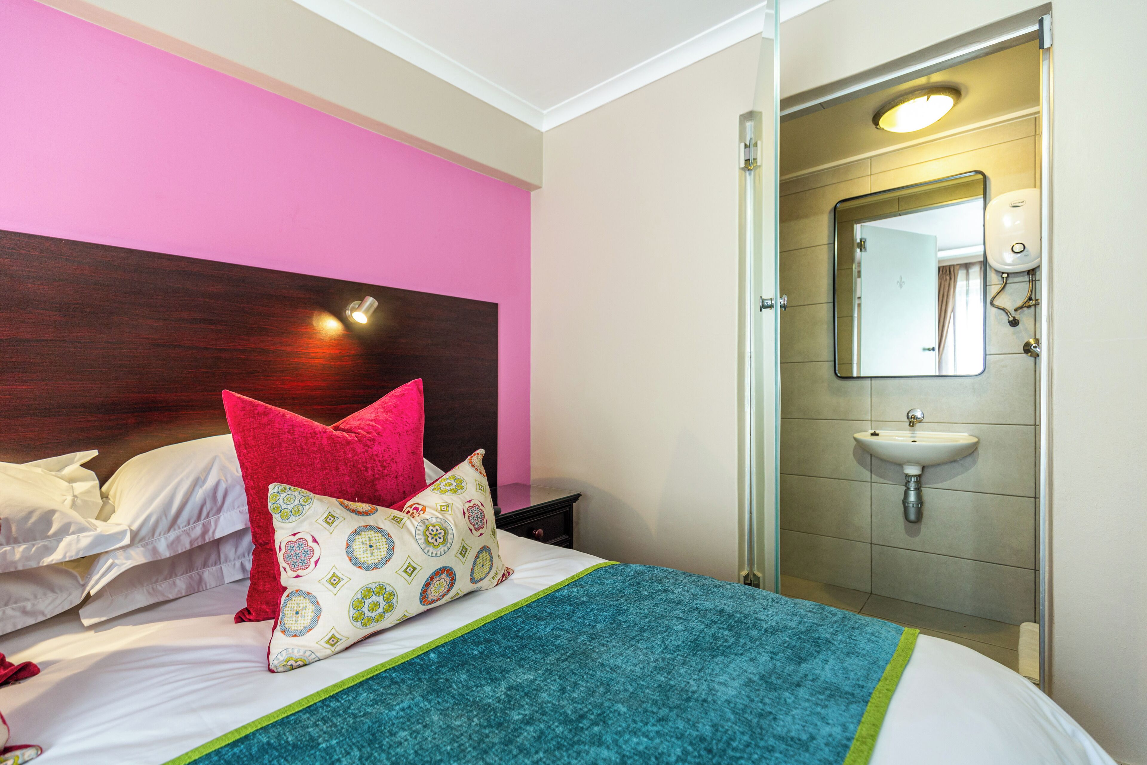Una habitación de hotel con cama, almohadas, mesita de noche y baño con lavamanos y espejo.