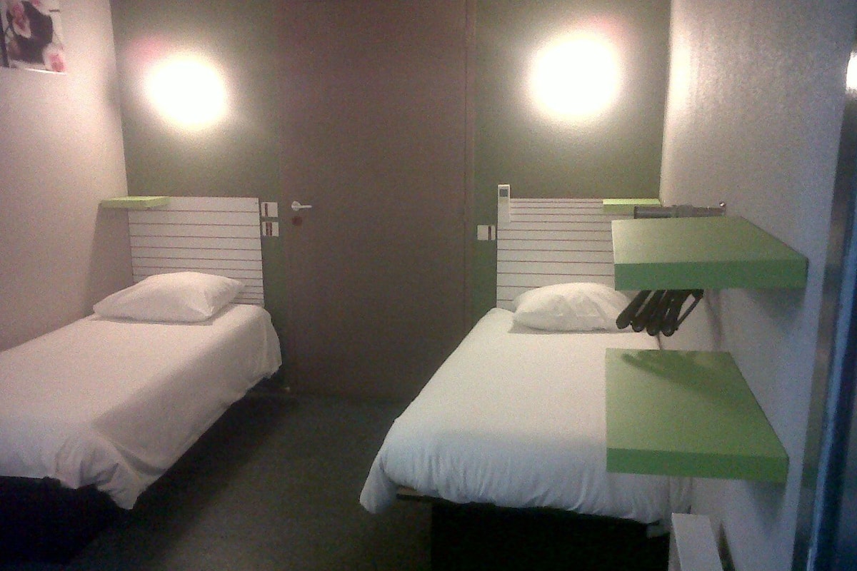 A small, dimly lit room with two single beds, each with a pillow and a green shelf.