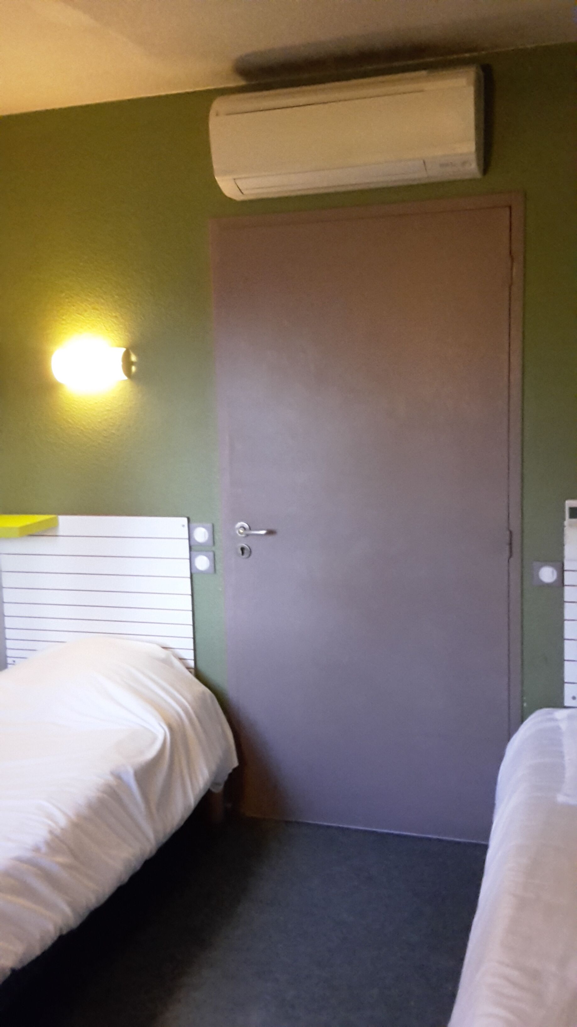 Une chambre d’hôtel avec deux lits, une porte, un climatiseur fixé au mur et un luminaire.