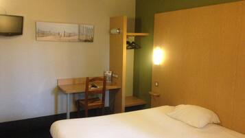 Una habitación de hotel con cama, escritorio, silla, televisión y lámpara fijada a la pared.