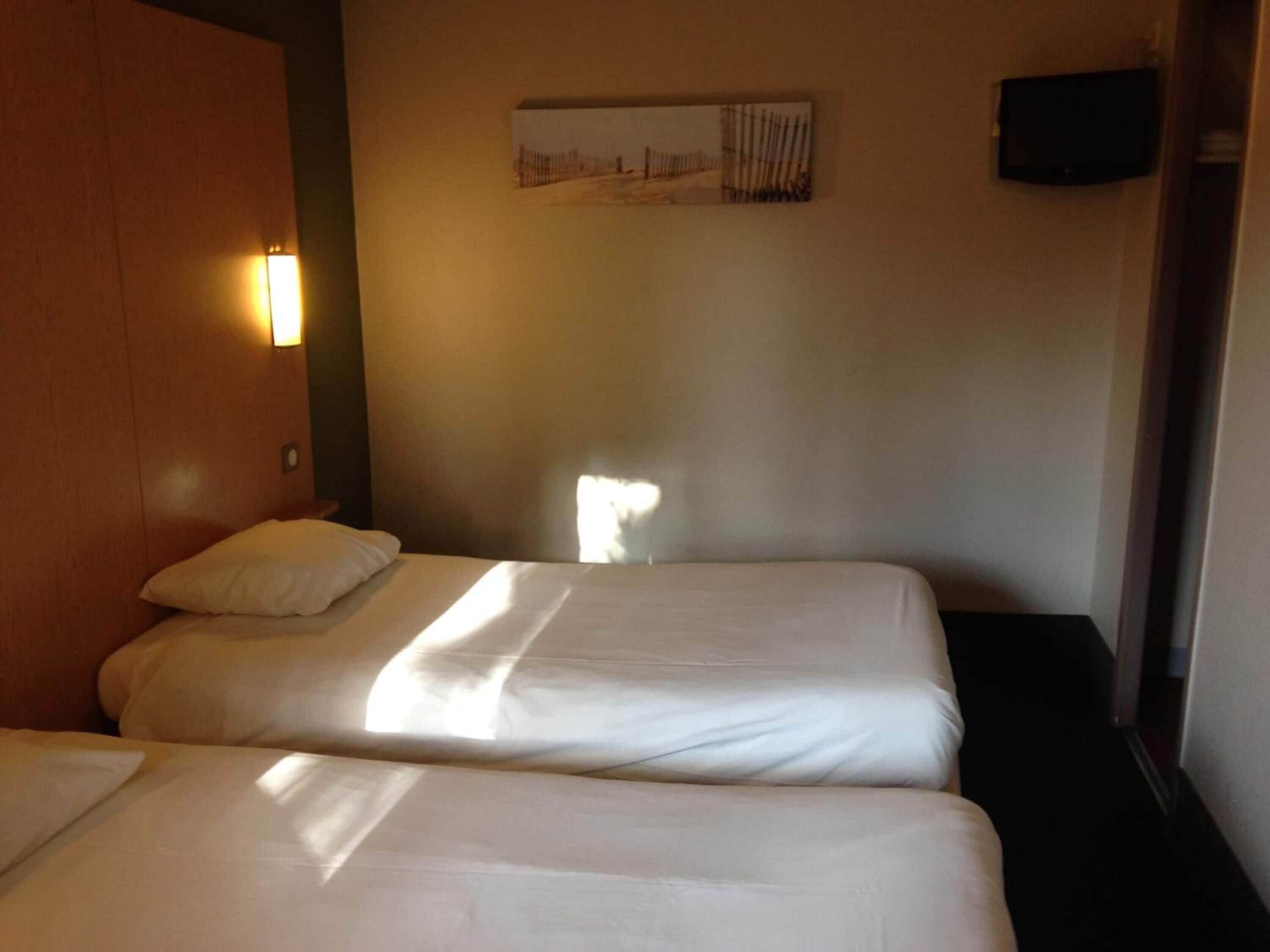 Habitación de hotel con dos camas, un televisor instalado en la pared y una ventana que deja entrar luz natural.