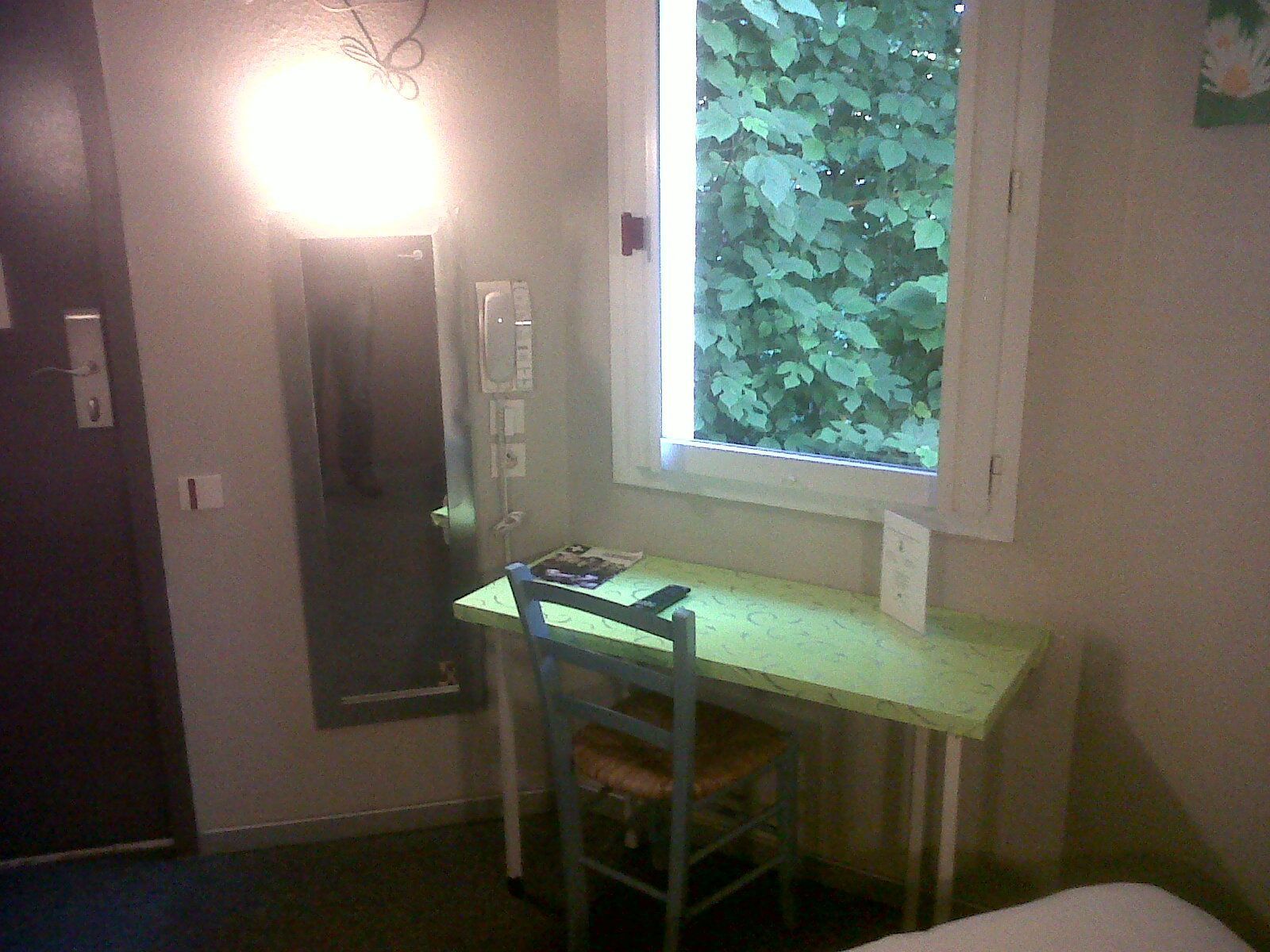 Une petite pièce avec un bureau et une chaise, une fenêtre donnant sur la verdure et une porte.