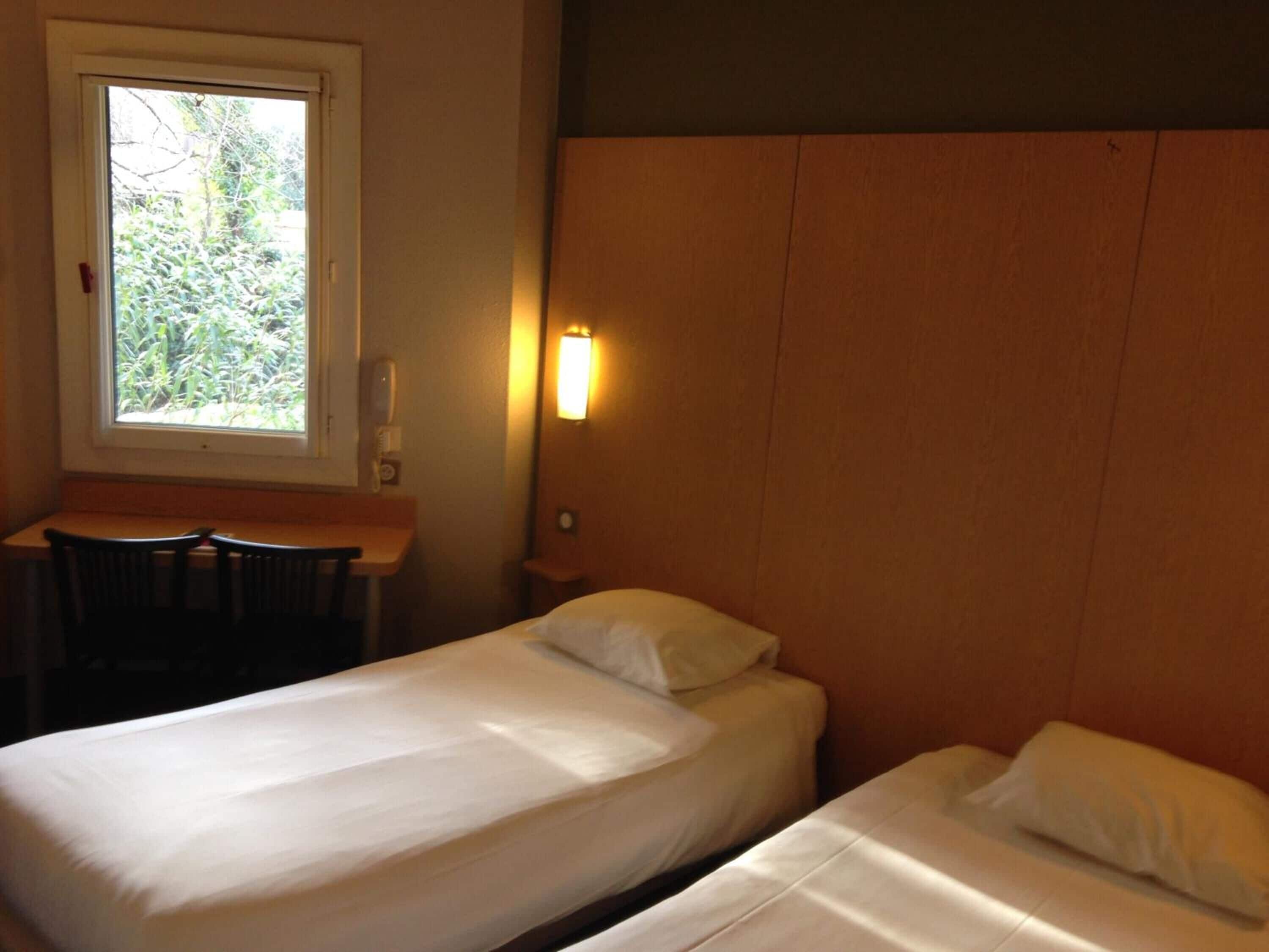 Een hotelkamer met twee bedden, een klein bureau en een raam met uitzicht op groen.