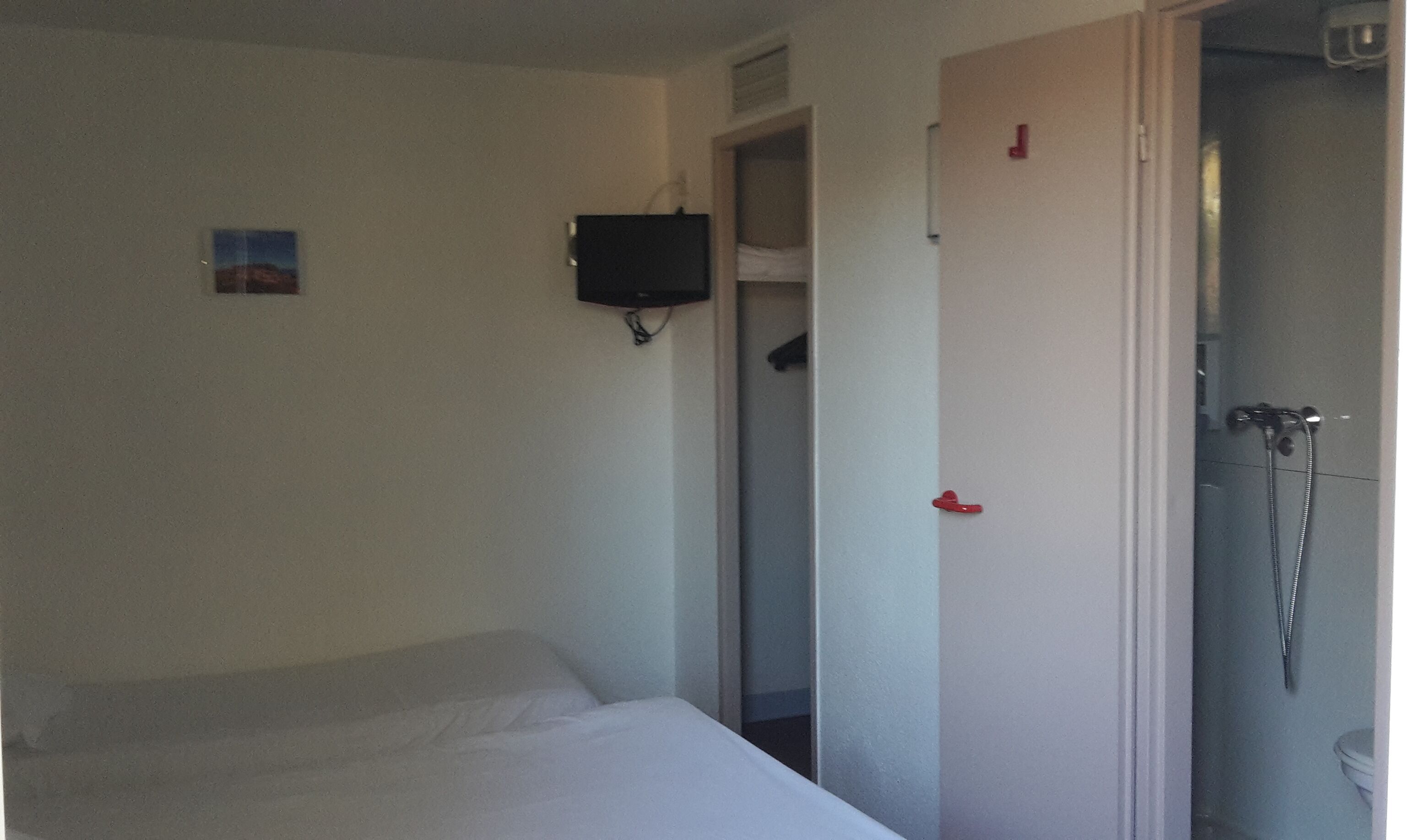 A small, sparsely furnished room with a bed, a television mounted on the wall, and a door leading to a bathroom with a shower.