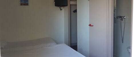 A small, sparsely furnished room with a bed, a television mounted on the wall, and a door leading to a bathroom with a shower.