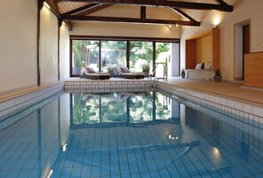 Una piscina interior con suelo de baldosas, un área de descanso con asientos y grandes ventanales que permiten la entrada de luz natural.