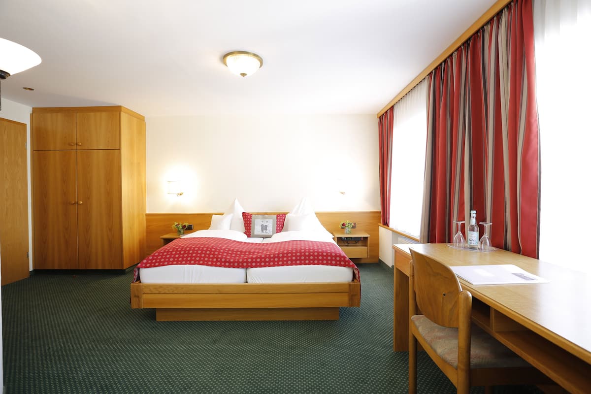 Una habitación de hotel con cama, escritorio, silla, armario y mesita de noche con lámpara.
