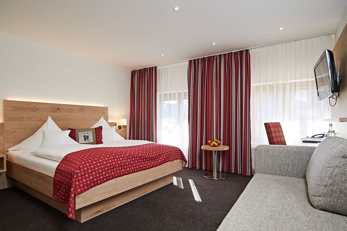 Habitación de hotel con una cama de madera, un sofá gris, una mesita redonda con una cesta de frutas y un televisor en la pared.